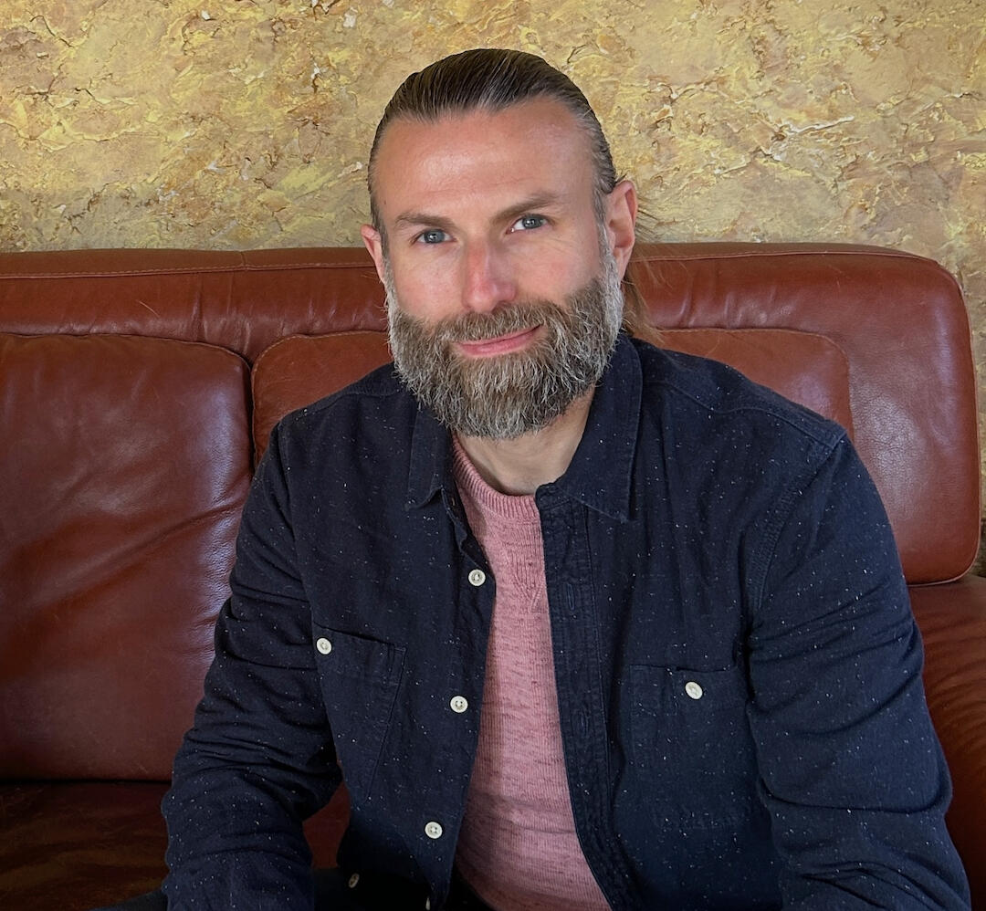 Jake Jones Jake Jones Headshot sitting on brown couch with navy blue shirt and red sweater